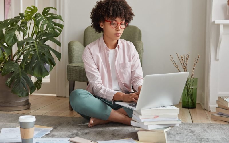Black businesswoman keyboards on laptop computer, makes diploma paper, uses laptop which stands on pile of books, drinks takeaway coffee, sits crossed legs on floor, busy working. Studying concept